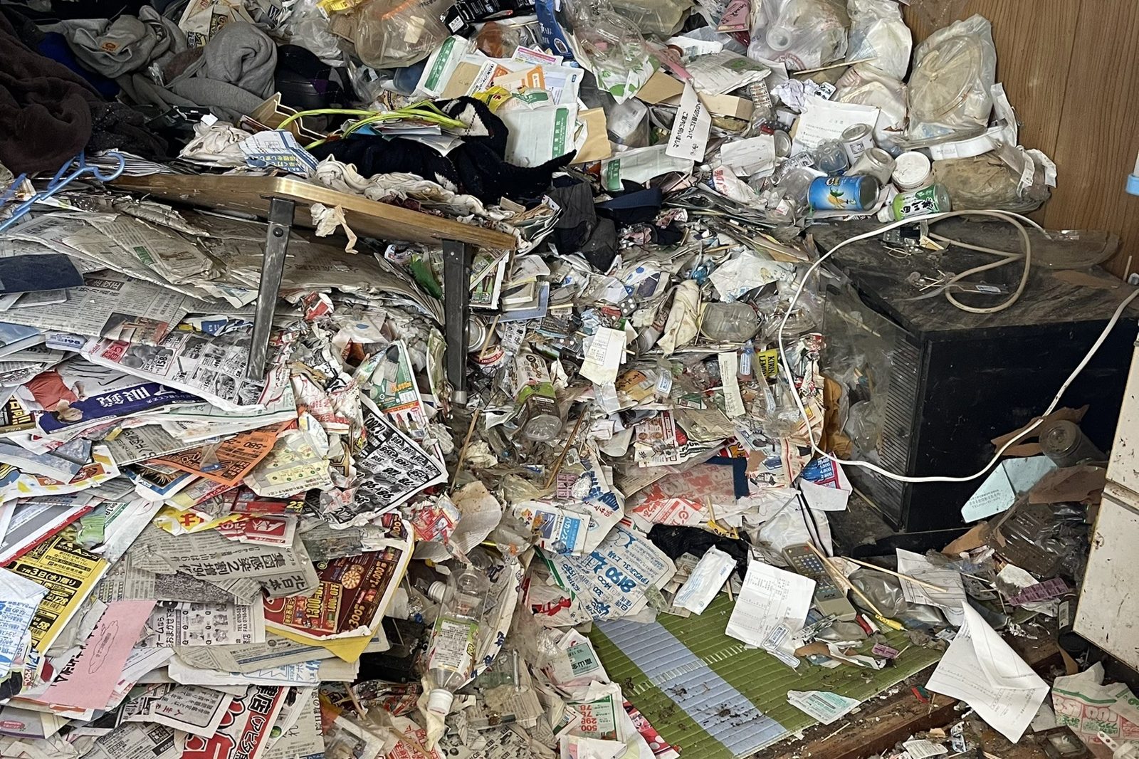 ゴミ屋敷の片付け前の部屋（新聞や袋ごみが積み重なった様子）
